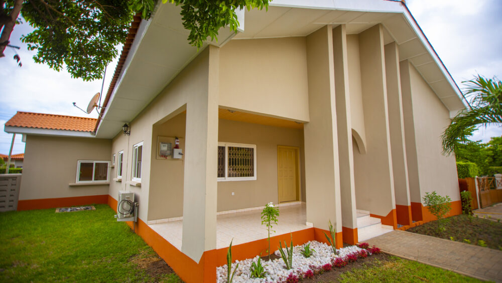 A 3 bedroom house painted in ivory colour and orange at the base with a garden surrounding it.