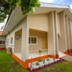 A 3 bedroom house painted in ivory colour and orange at the base with a garden surrounding it.