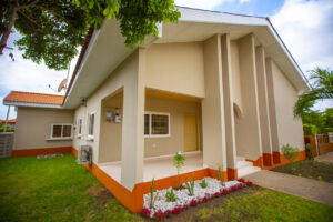 A 3 bedroom house painted in ivory colour and orange at the base with a garden surrounding it.