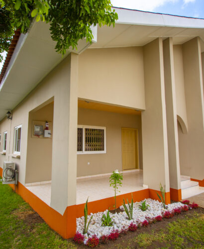 A 3 bedroom house painted in ivory colour and orange at the base with a garden surrounding it.